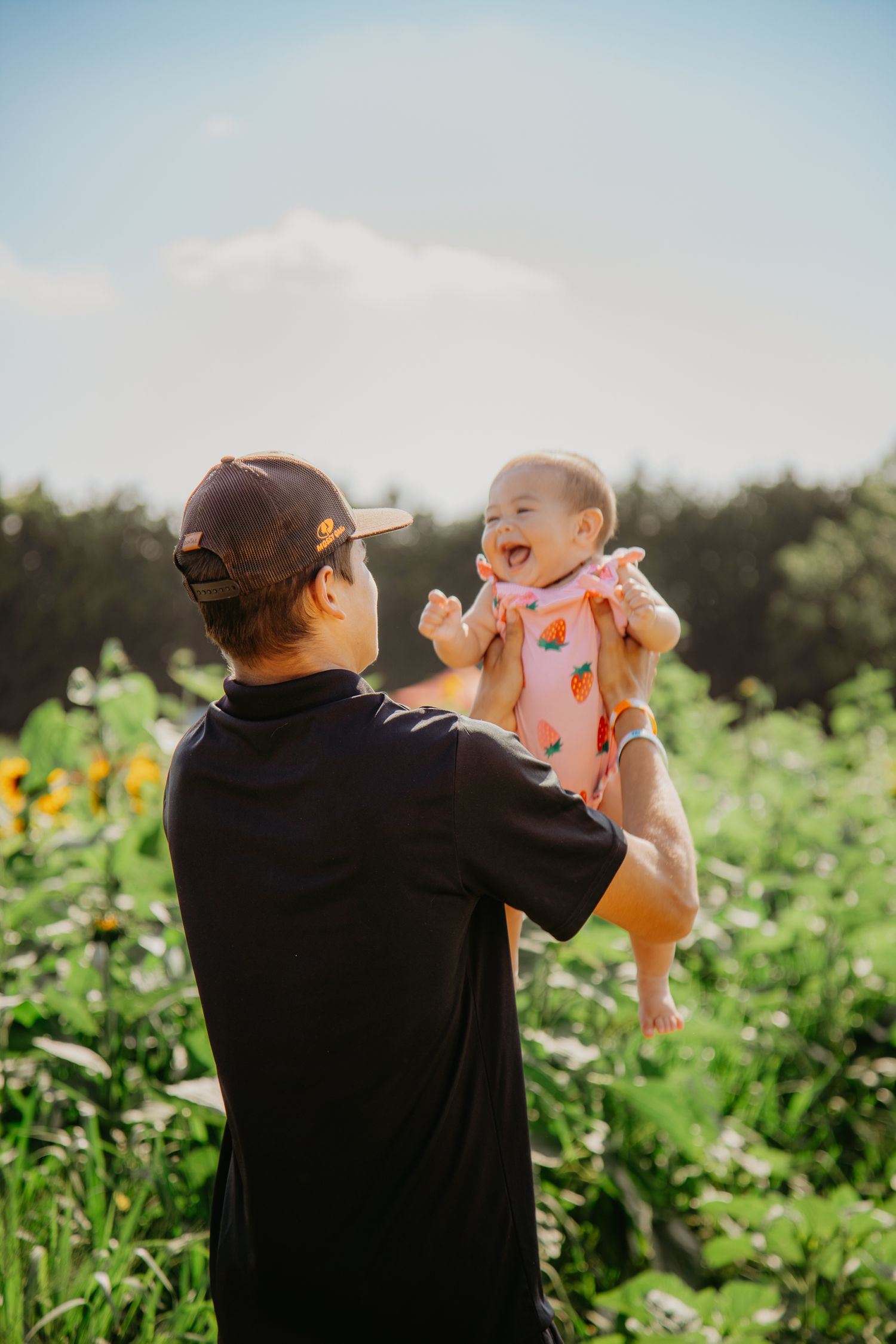 A joyful baby wearing pink polka dots is lifted high in a sunflower garden on a sunny summer day.