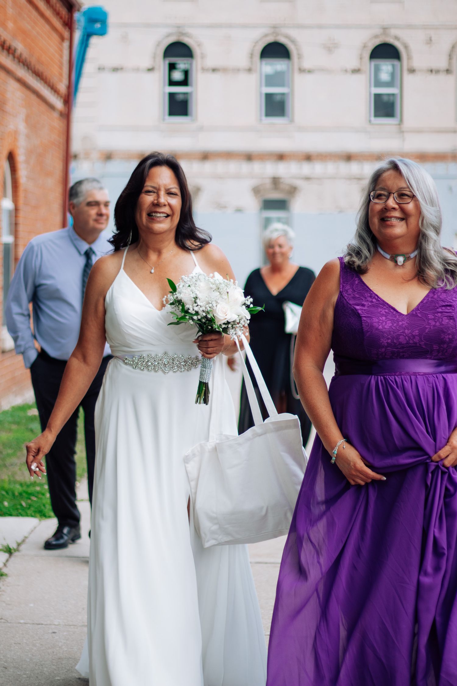 Bride in white gown holding bouquet walks with woman in purple dress on sidewalk outside brick building.