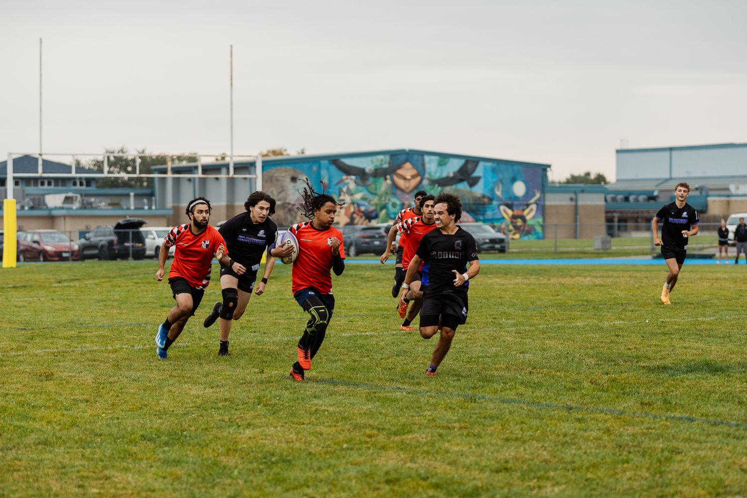 Rugby players sprint across a green field during an outdoor match, with a colorful mural and industrial buildings in the background.