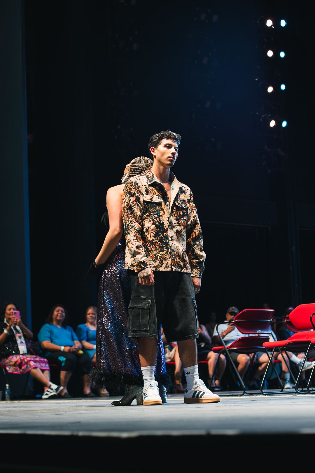 Male model in floral jacket and cargo shorts walks runway at fashion show with audience seated in background.