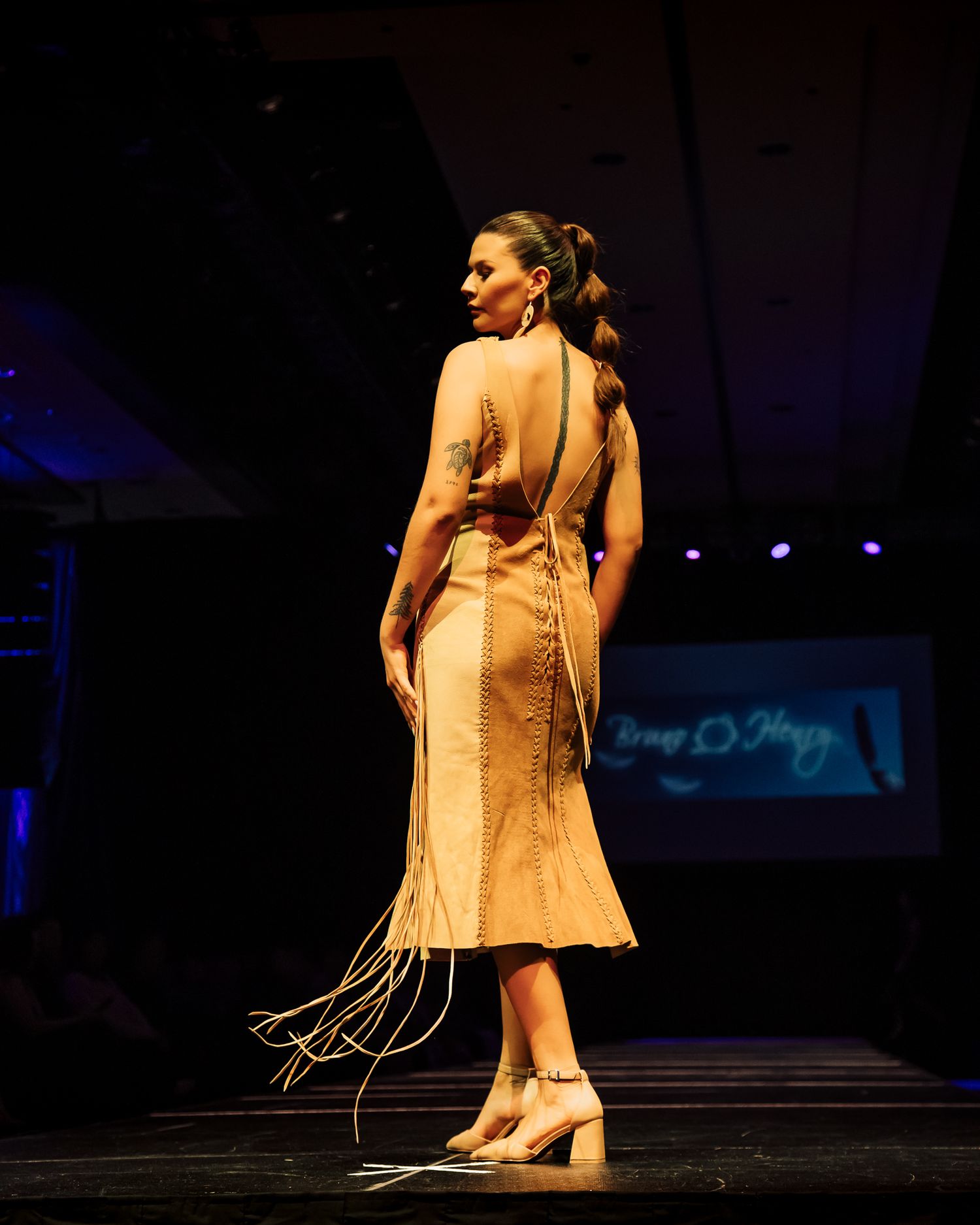 Model walks runway in backless tan fringe dress with braided hair at fashion show.