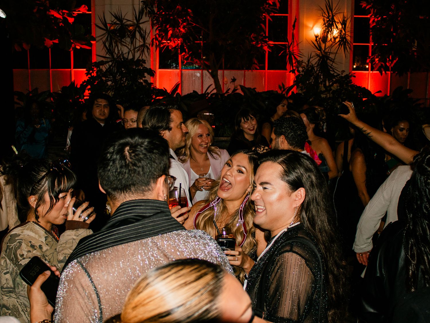 Crowd of elegantly dressed guests socializing at a glamorous indoor evening party with red lighting.