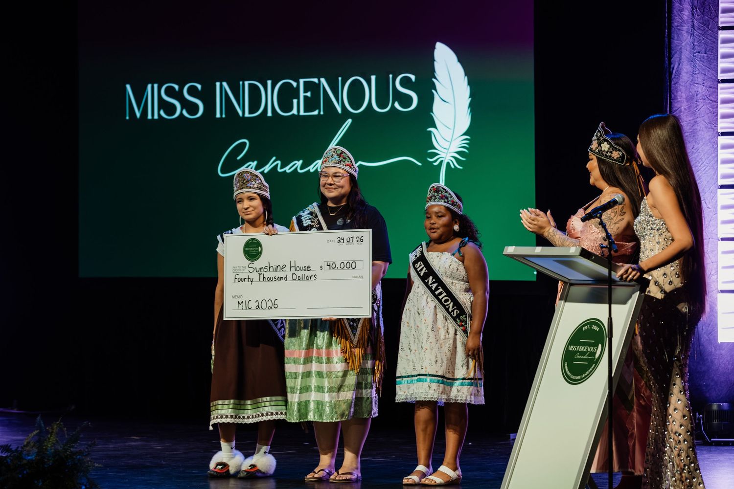 Miss Indigenous Canada pageant winner receives a large prize check on stage under the event logo.