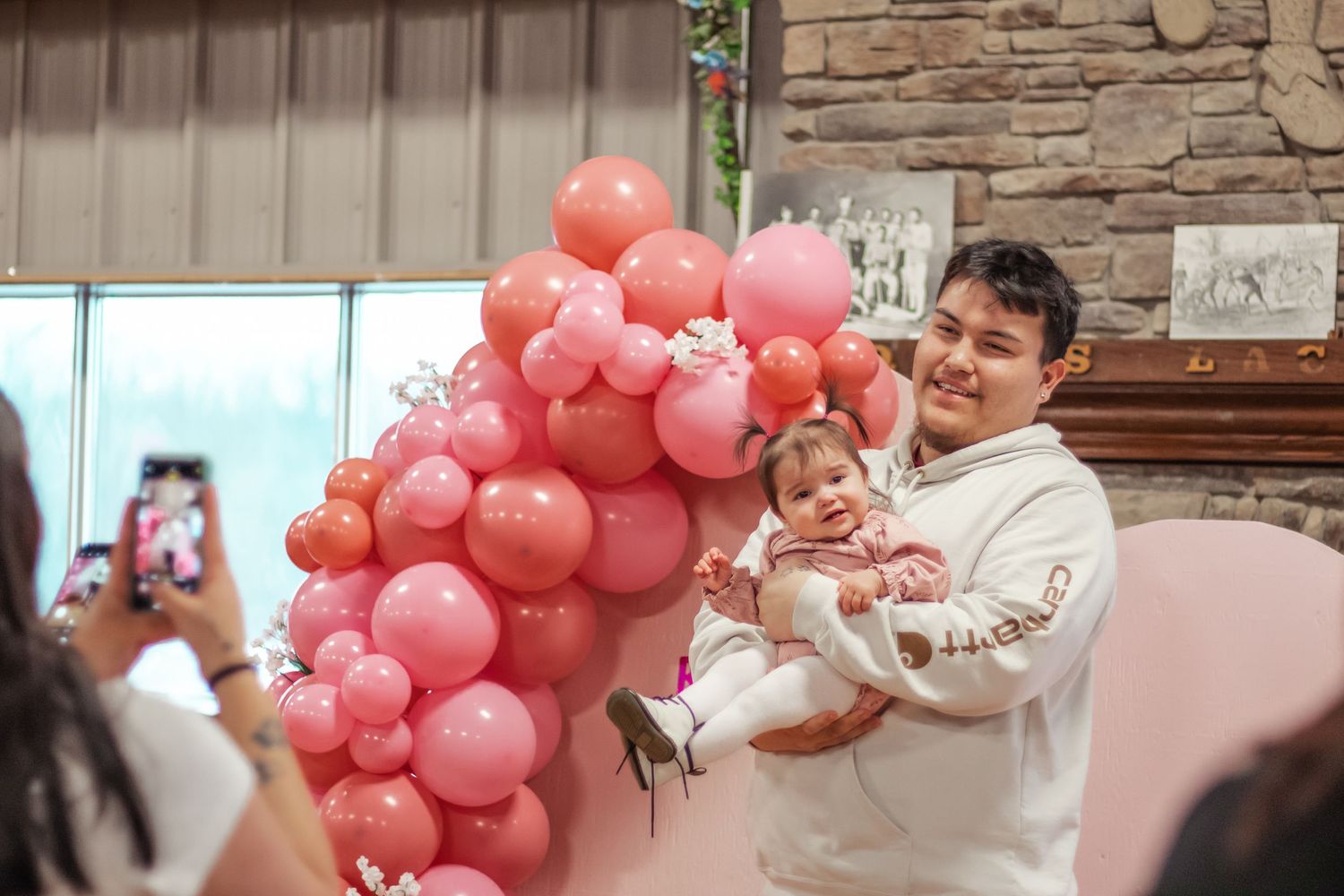 A man smiles holding a baby girl near a pink balloon arrangement at a birthday party celebration indoors.