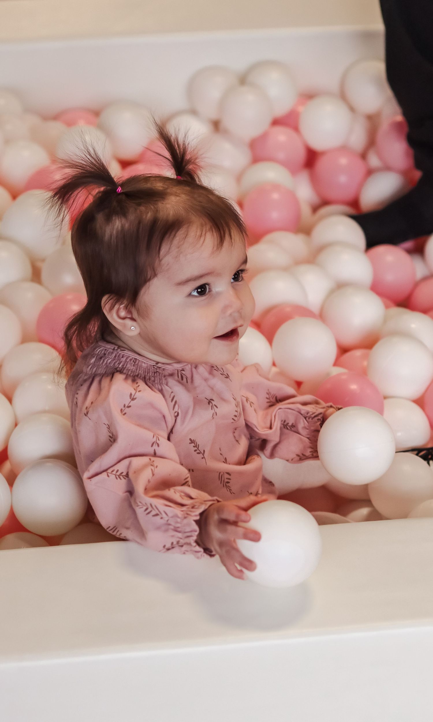 Adorable baby girl in pink outfit playing in a white ball pit filled with pink and white balls.