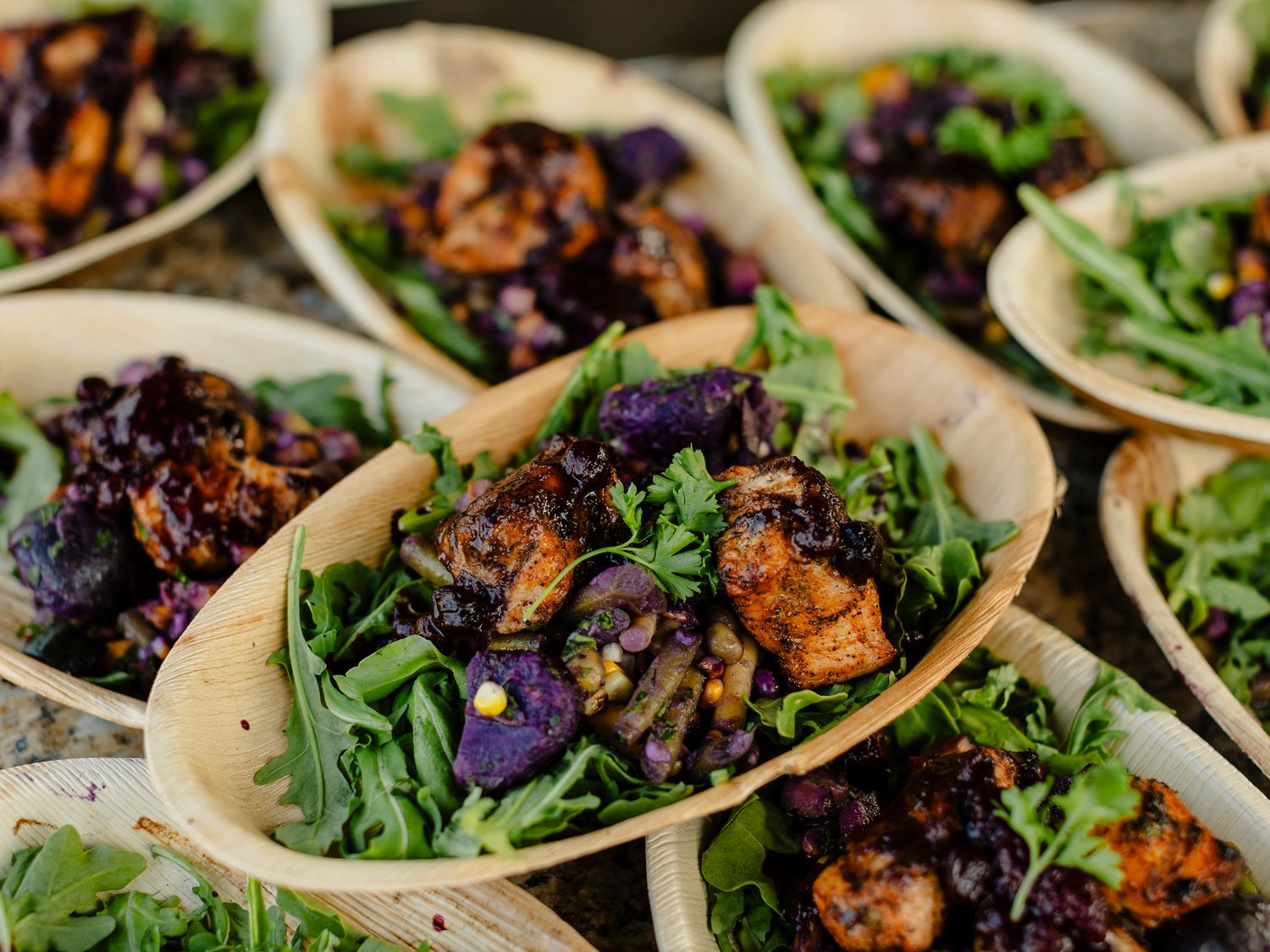 Wooden bowls filled with grilled chicken, black rice, purple potatoes, and fresh arugula salad at a catered event.