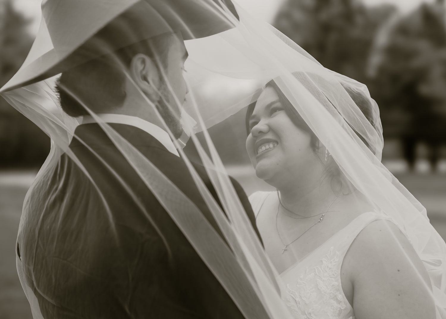 Black and white wedding photo of bride and groom sharing intimate moment under flowing veil outdoors.