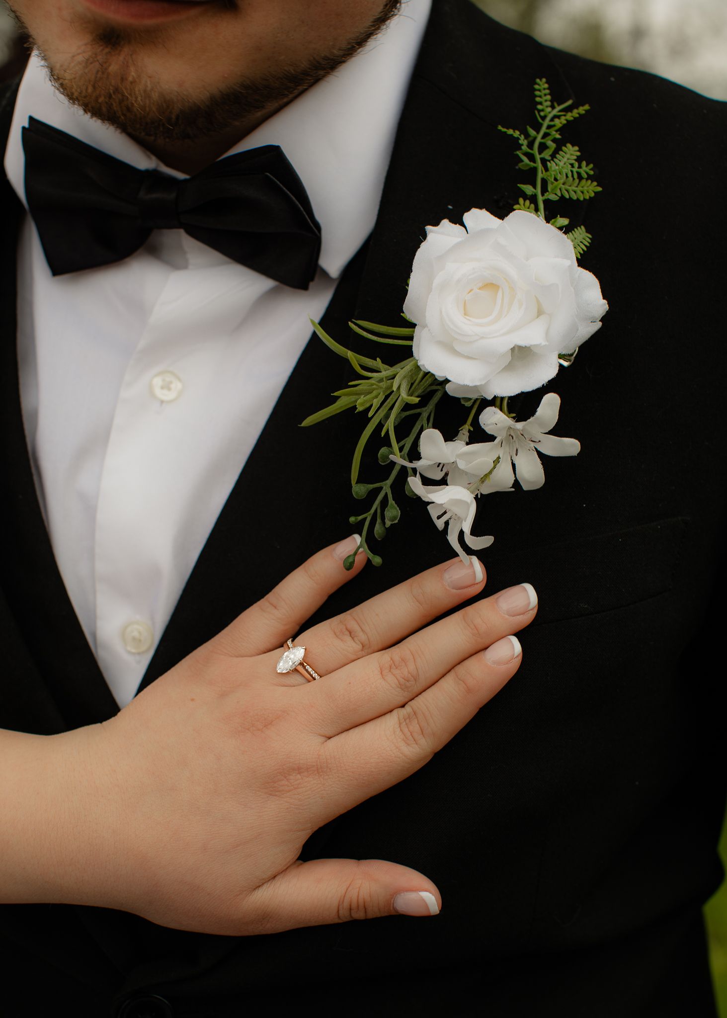 Groom in black tuxedo with white rose boutonniere as bride places hand on chest showing engagement ring.