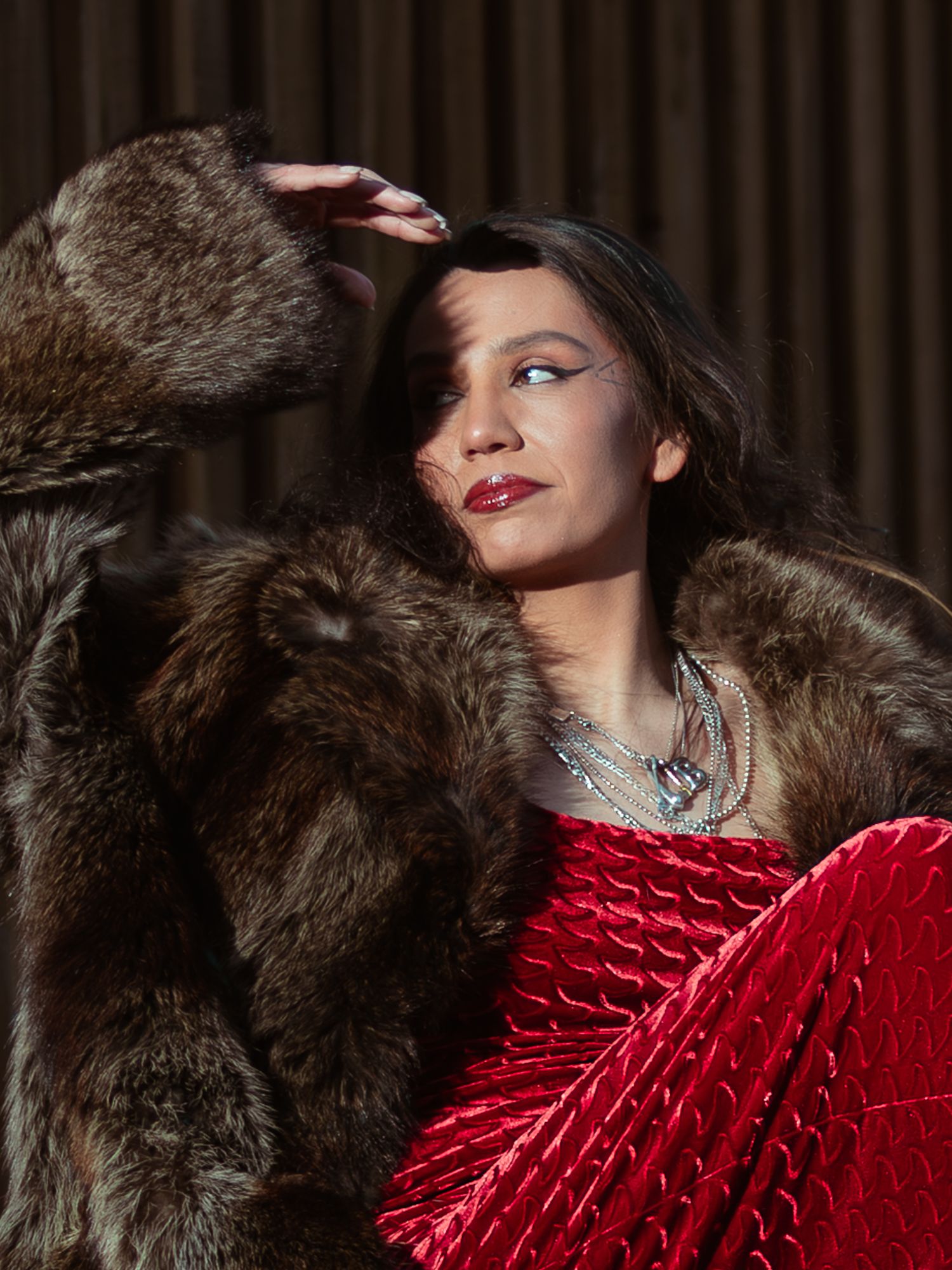 Woman in red velvet dress and brown fur coat with silver necklace in dramatic lighting.