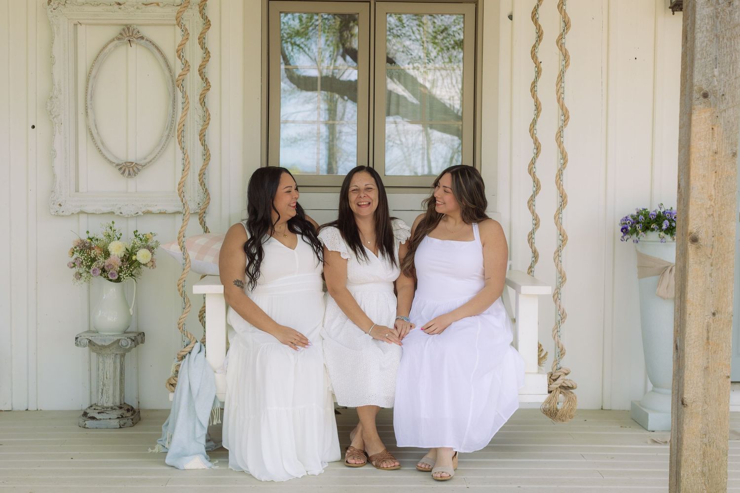 Three women in white dresses pose together on a rustic porch, smiling warmly in an elegant outdoor setting.
