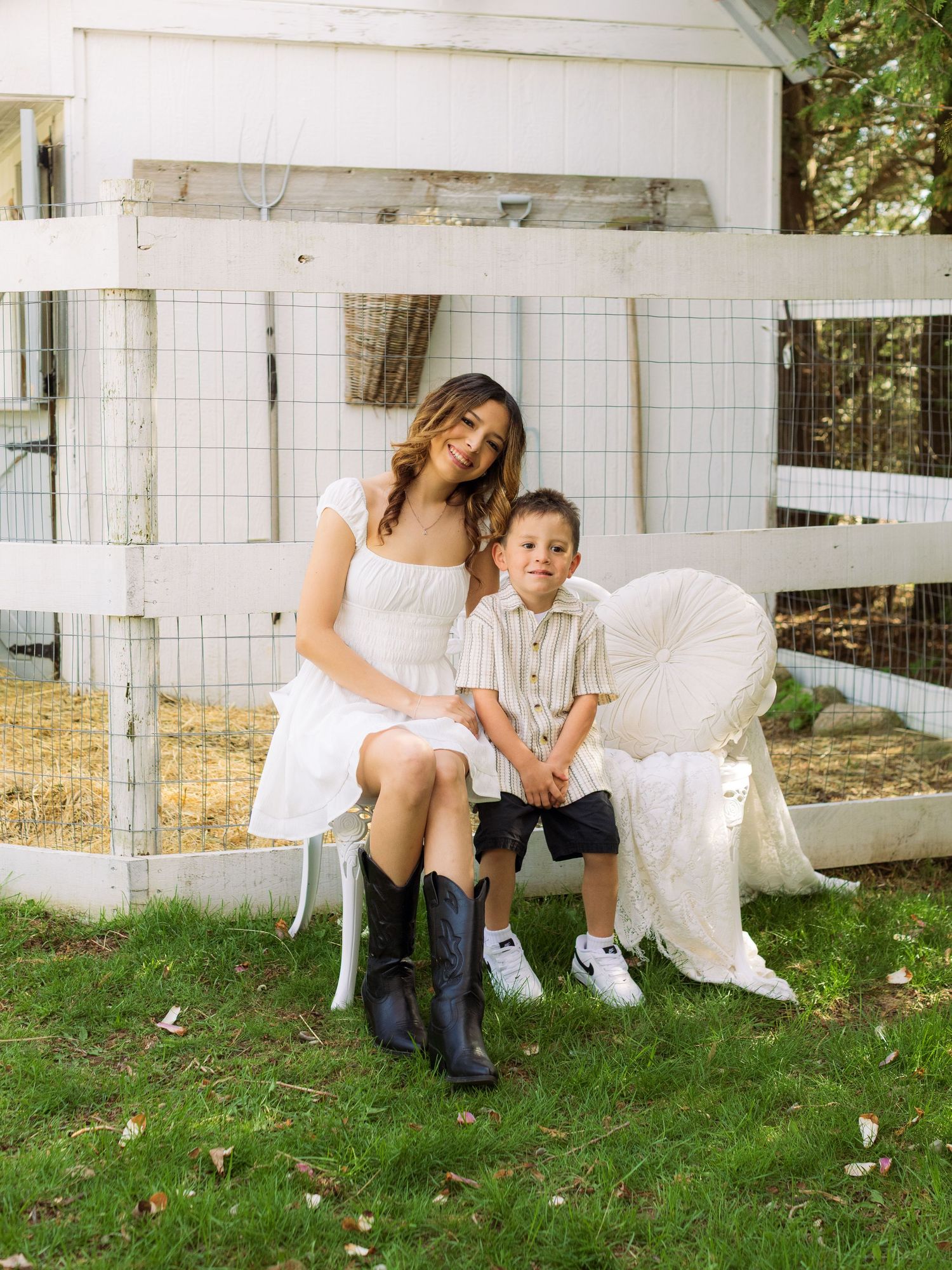 Woman in white dress with black boots and young boy pose by white fence in sunny backyard.