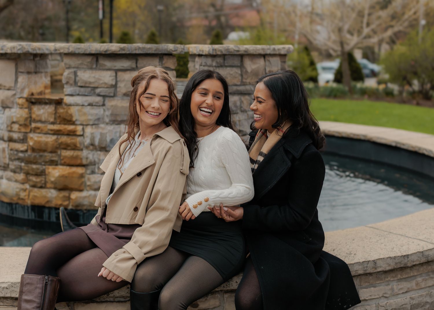 Three women laughing together by a stone fountain outdoors.