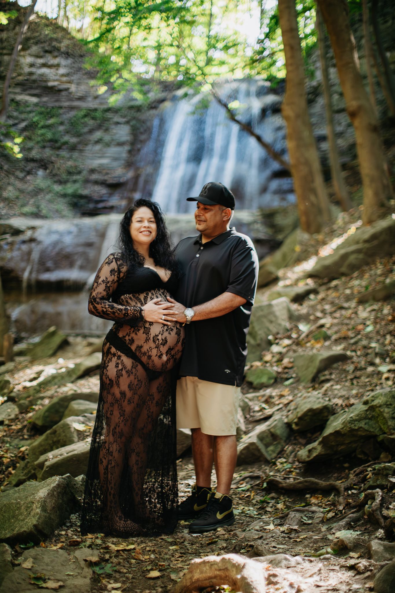 A romantic maternity photoshoot in front of a scenic waterfall with a couple embracing in nature.