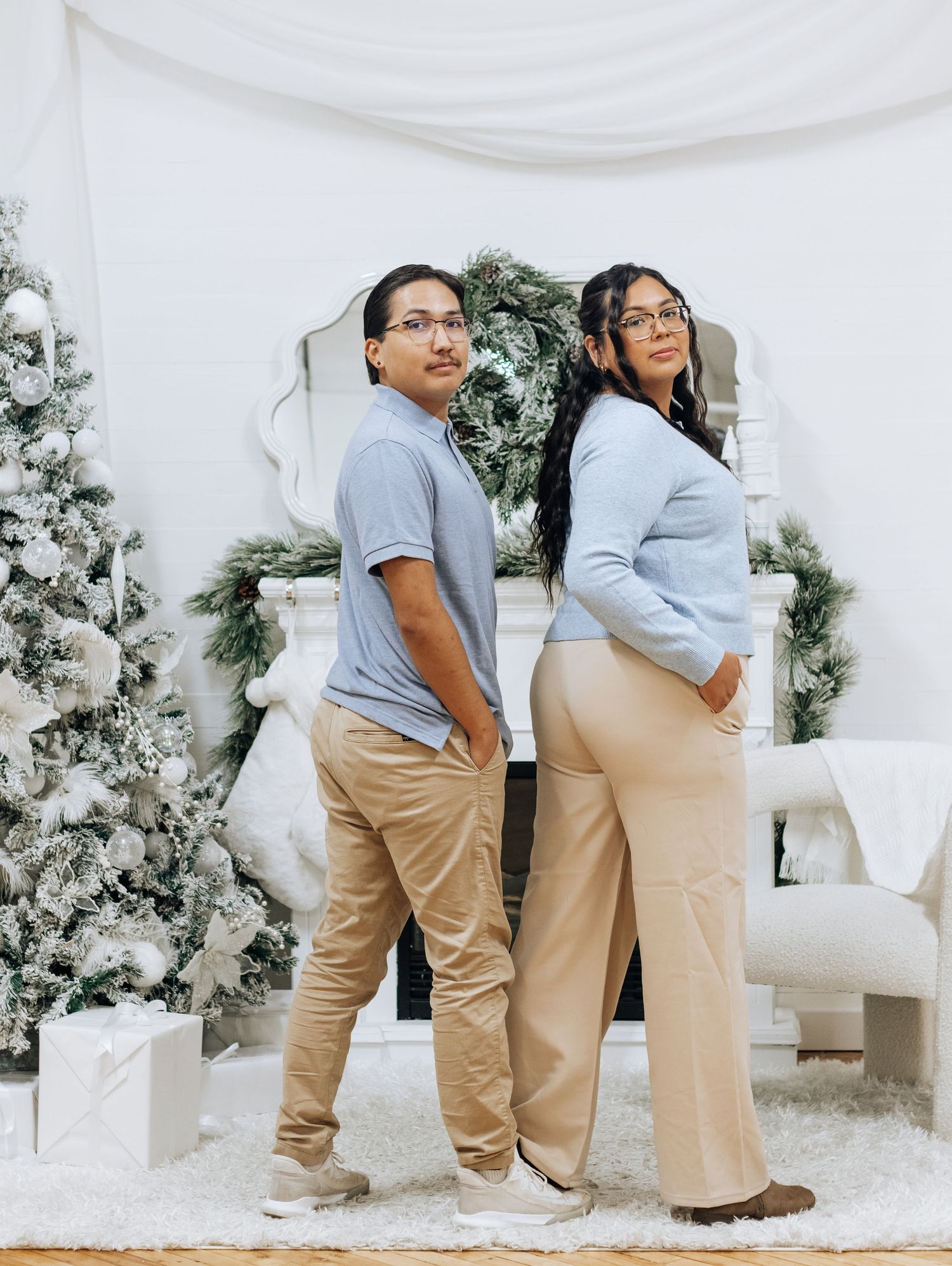 Two people in matching gray shirts and khaki pants pose playfully in a white winter-themed studio setting.
