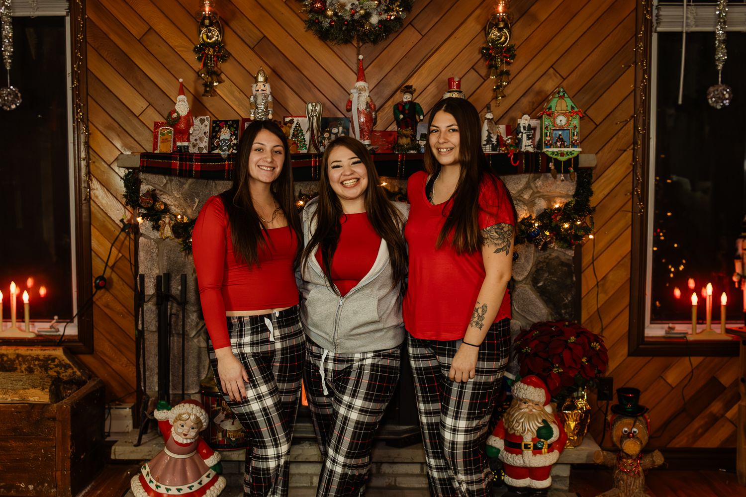Three people in matching red tops and plaid pants pose in front of rustic Christmas backdrop.