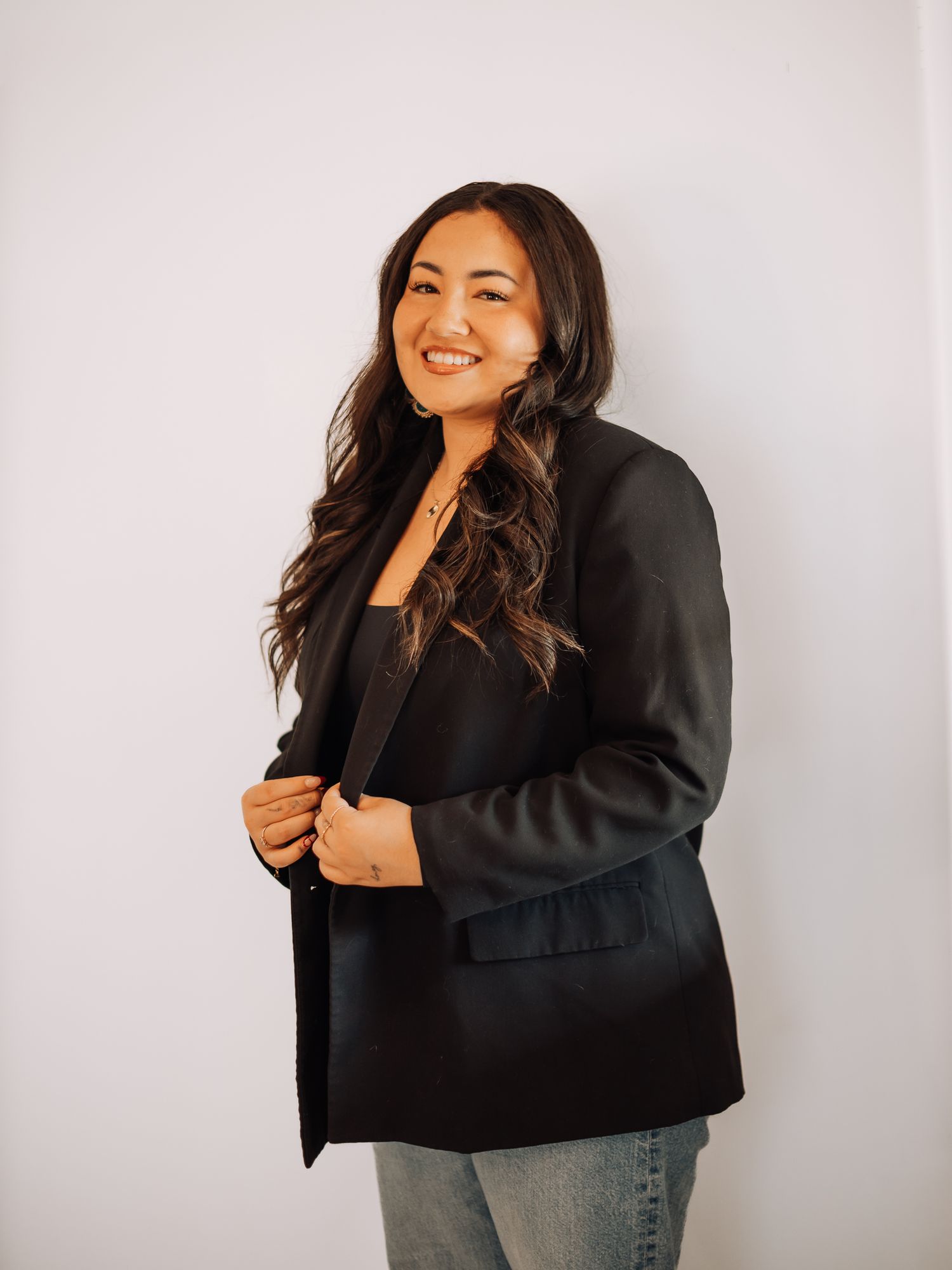A person wearing a black blazer and jeans poses casually against a white wall.