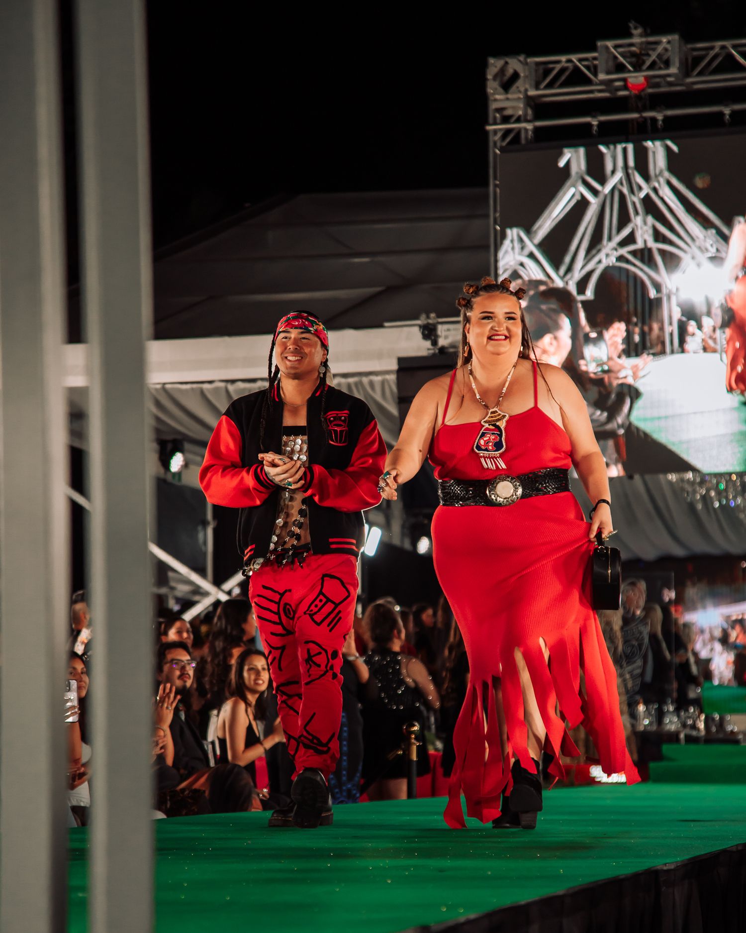 Models in vibrant red dresses perform choreographed movements on a green runway stage.