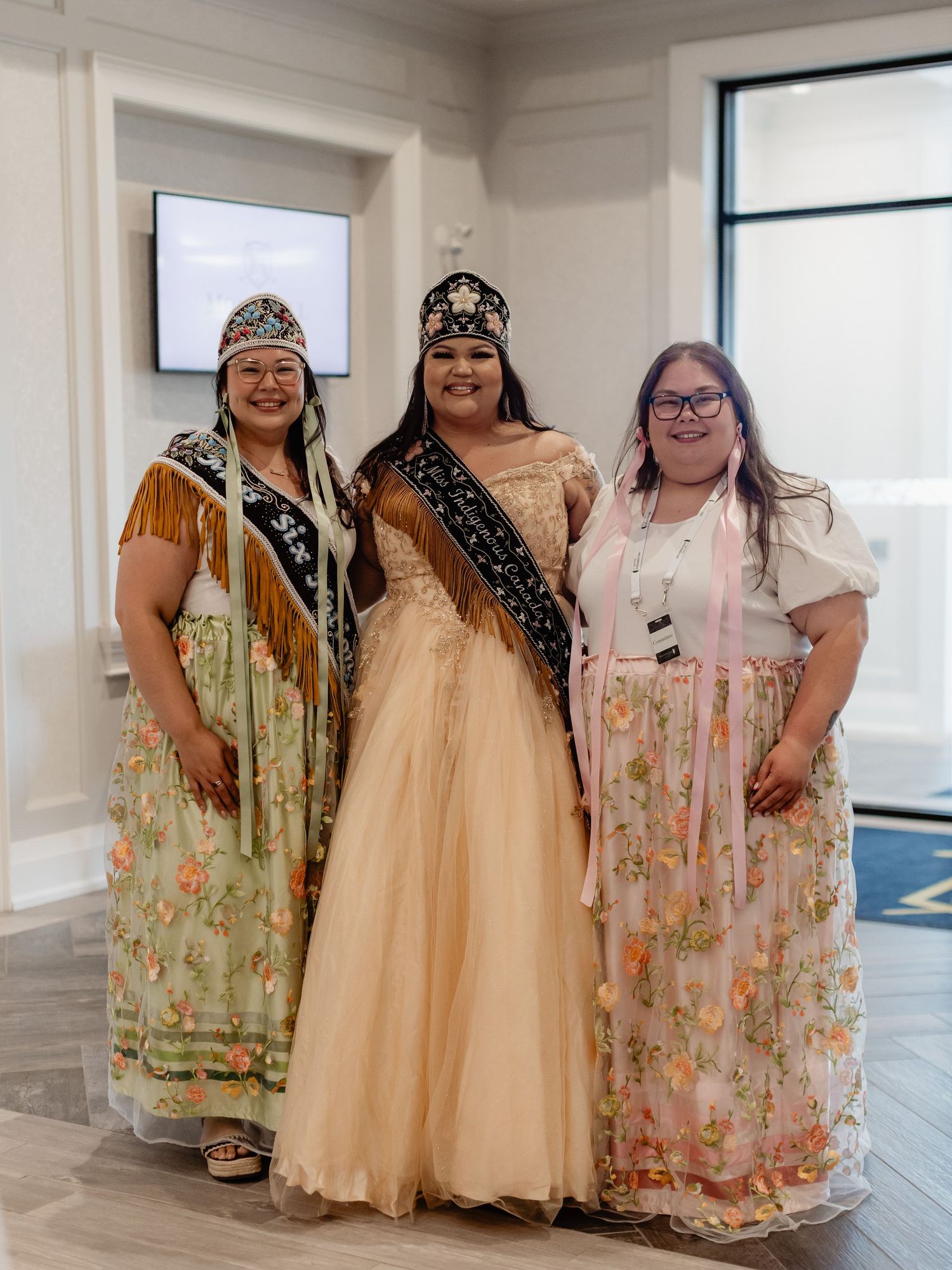 Three women in traditional Native American regalia and floral dresses pose together, with center woman wearing crown and sash.