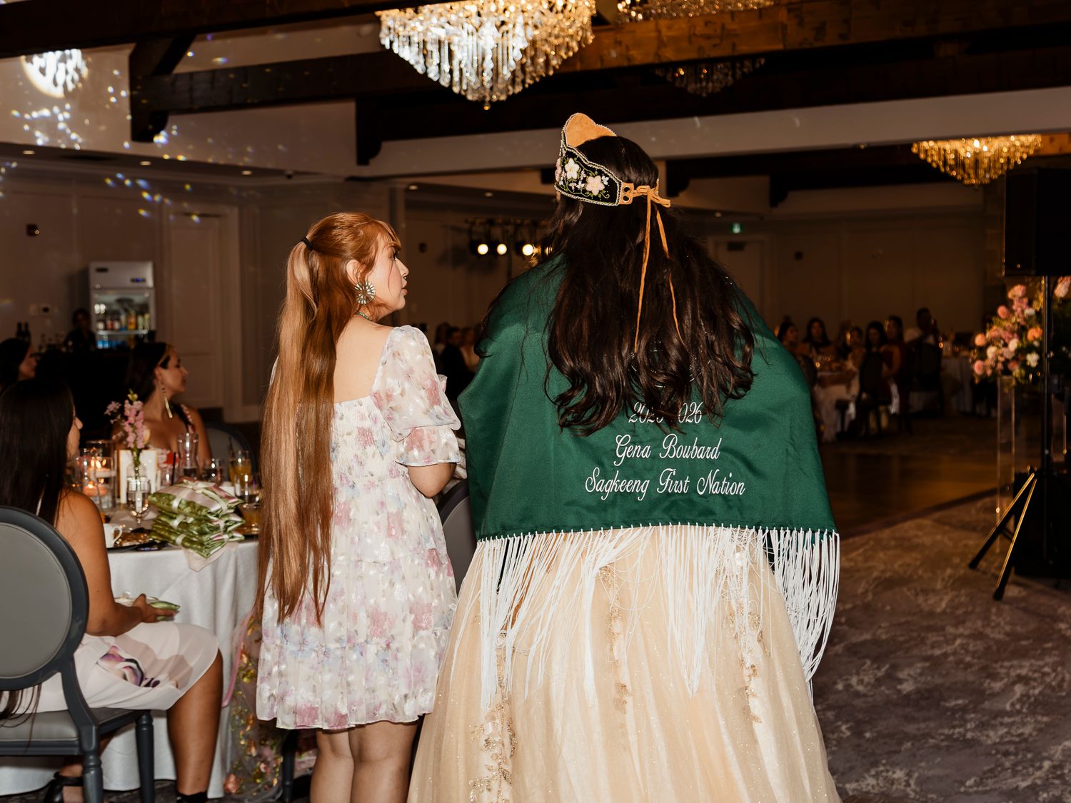 Two women at a formal banquet, one wearing a green embroidered jacket with fringe detailing at an elegant venue.