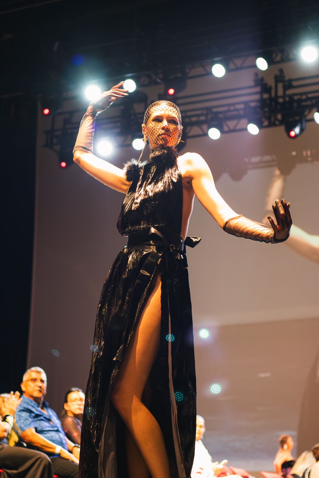 Model poses on runway in black halter gown with high slit at fashion show.