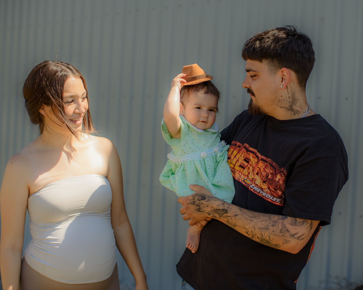Pregnant woman and tattooed man holding a baby wearing an orange hat outdoors near a corrugated wall.