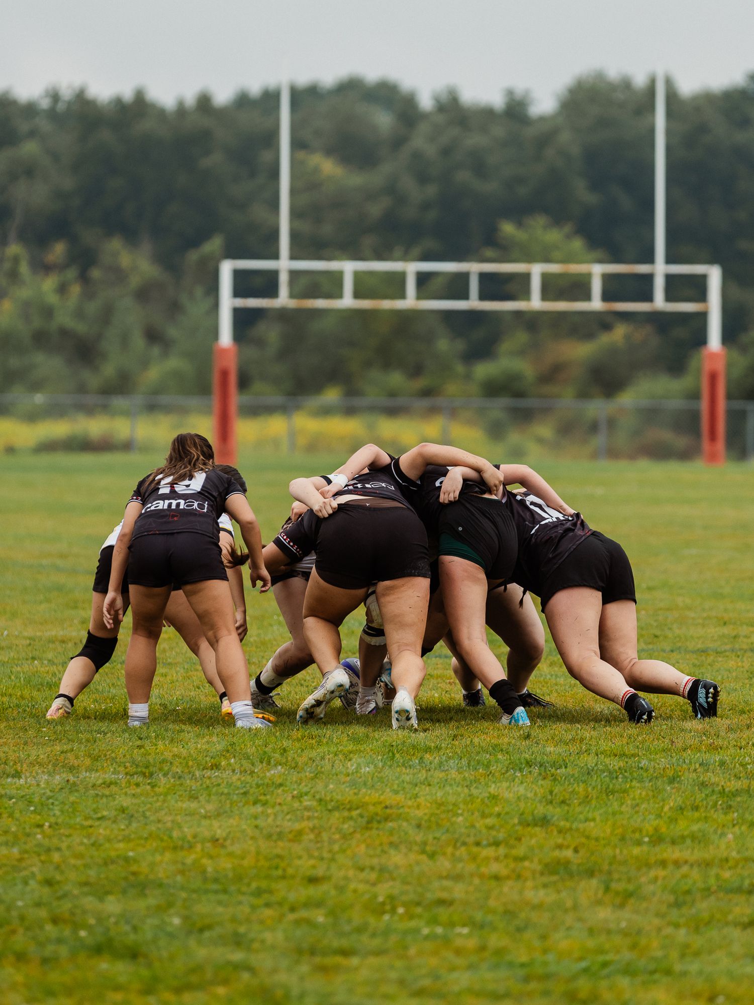Women rugby players in black uniforms form a scrum on green field with goalposts visible in background.