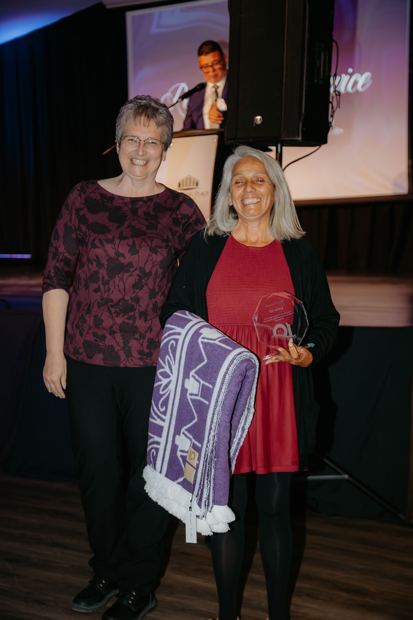 Two women pose together at awards ceremony, one holding glass trophy and purple blanket at indoor event.