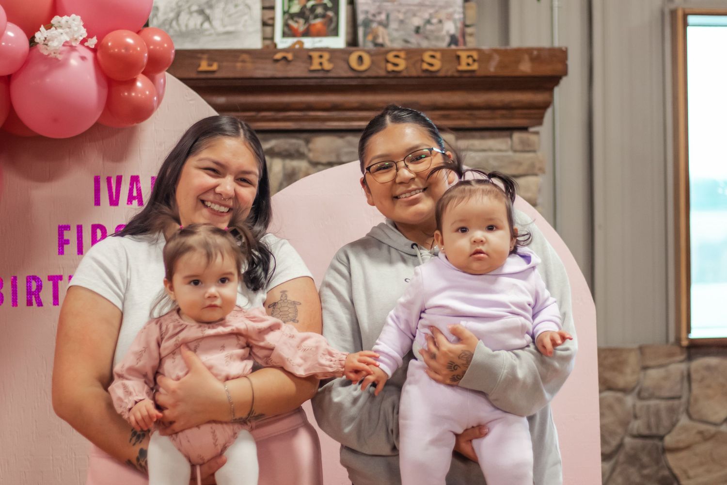 Two women holding babies at a first birthday party with pink balloons and decorative arch backdrop.