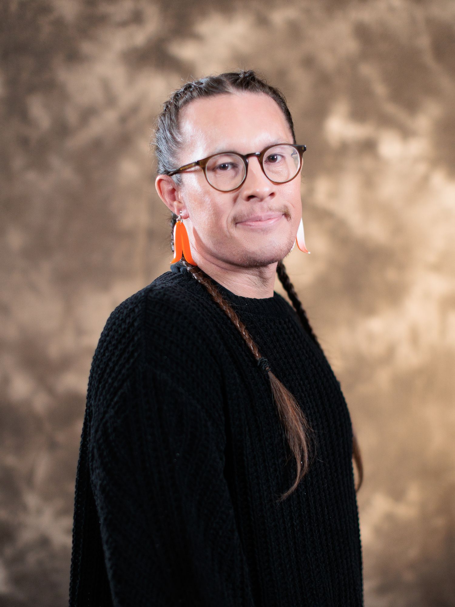 Person with braided hair, orange feather earrings, and glasses wearing a black cardigan against a brown backdrop.