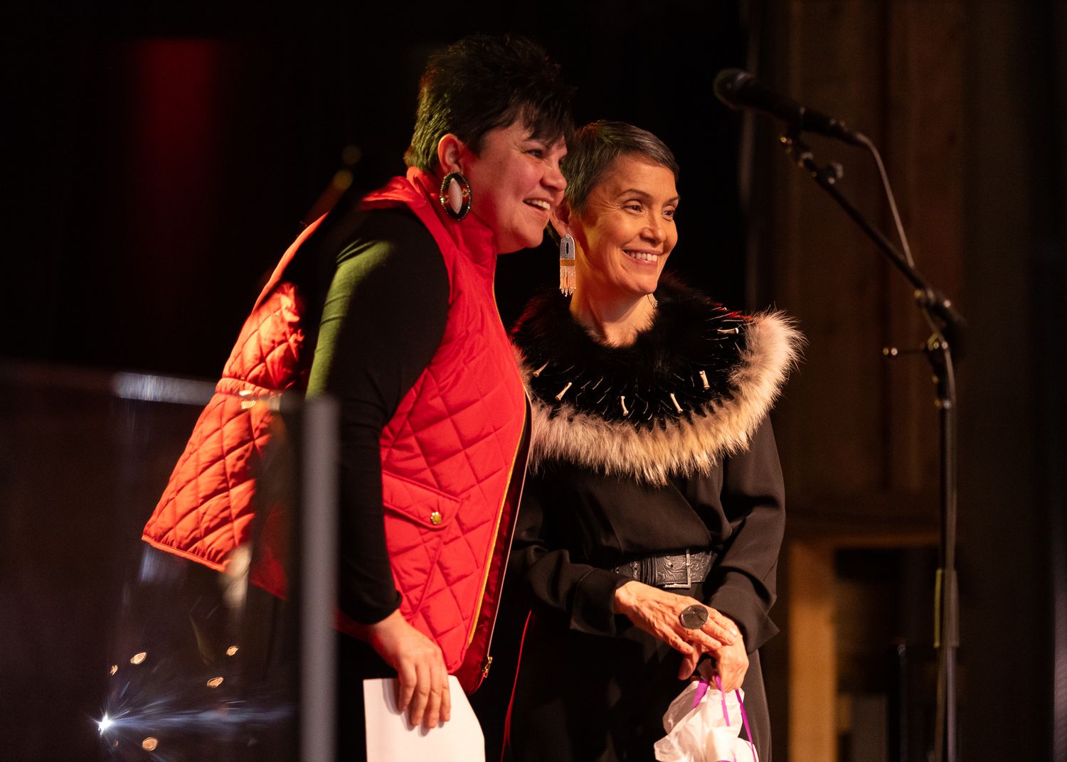 Two women smiling on stage, one in a red quilted vest and one in a black fur-trimmed jacket, at an indoor event.