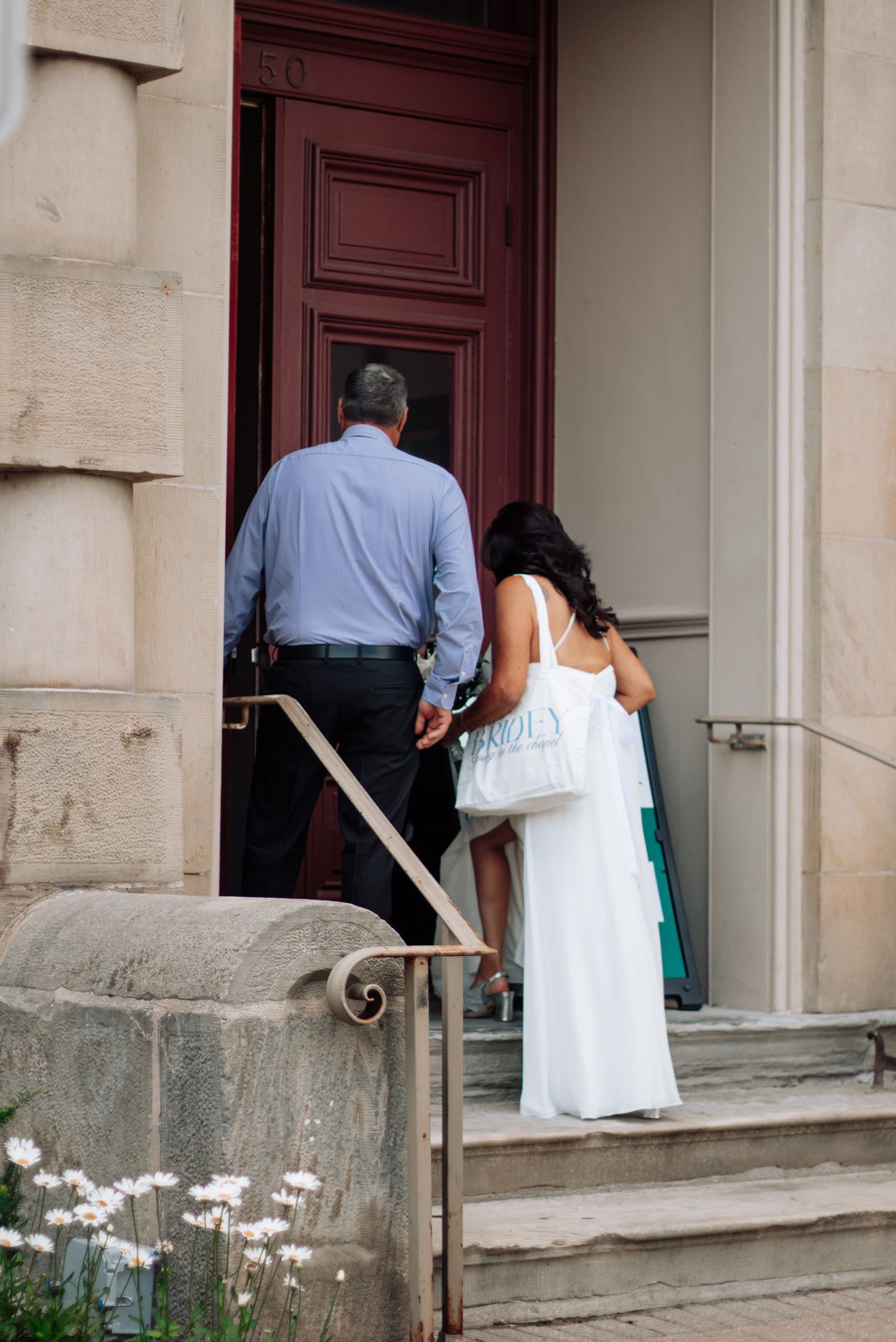 Bride in white gown and groom in blue shirt entering church through red doors on wedding day.