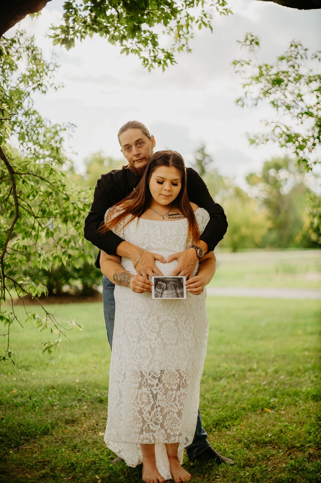 A couple poses together under tree branches while holding an ultrasound photo.