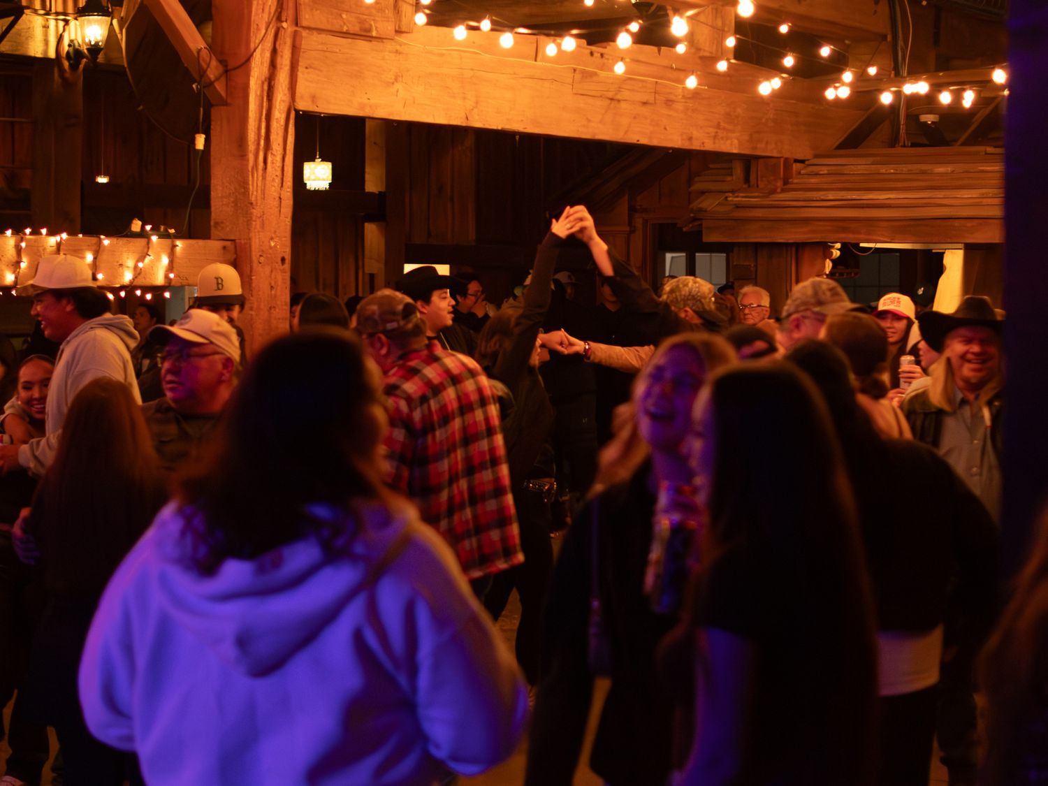 A crowd dances under string lights at a rustic indoor venue.