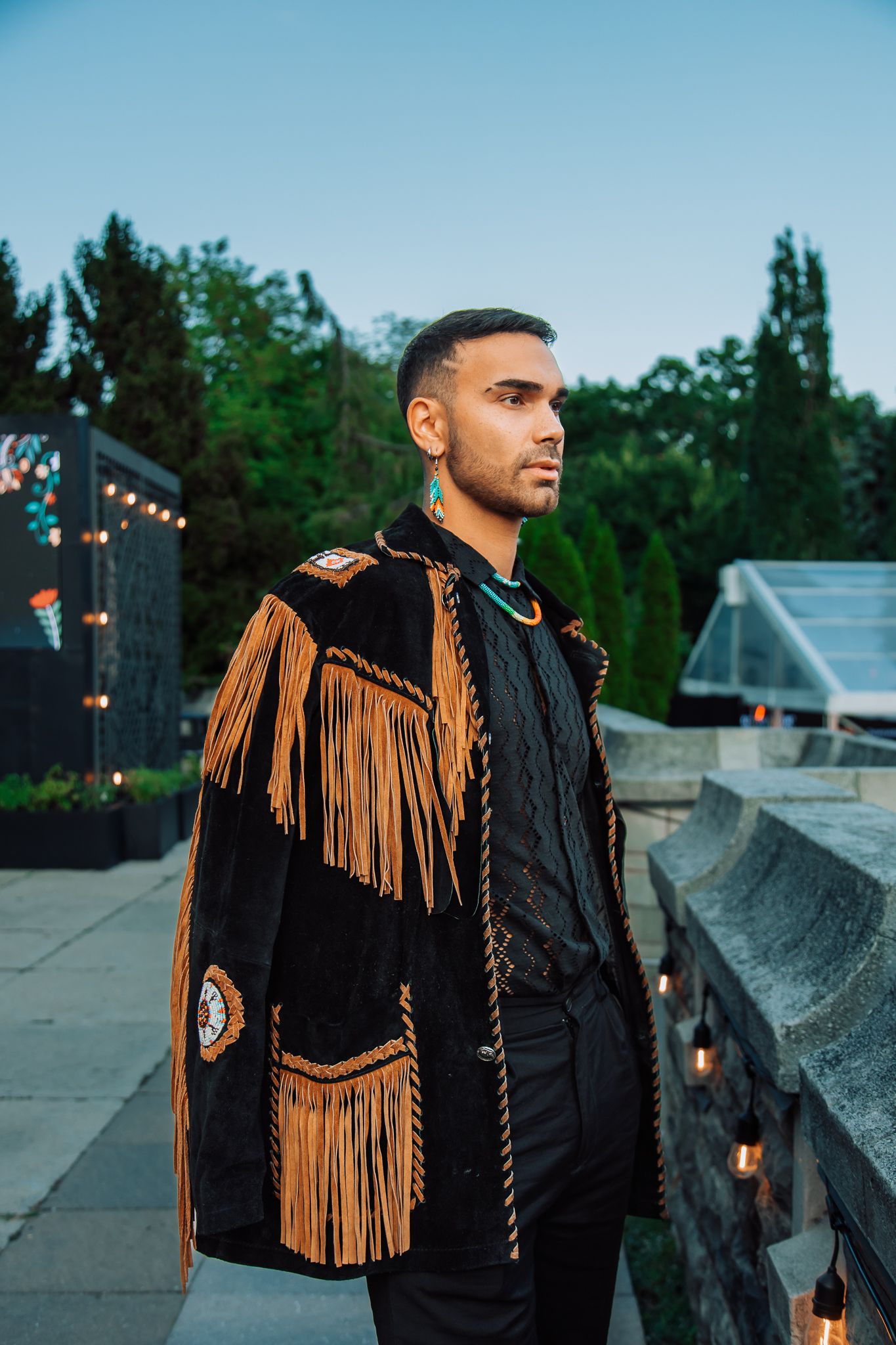 Person in black jacket with brown fringe detail posing against evening sky.