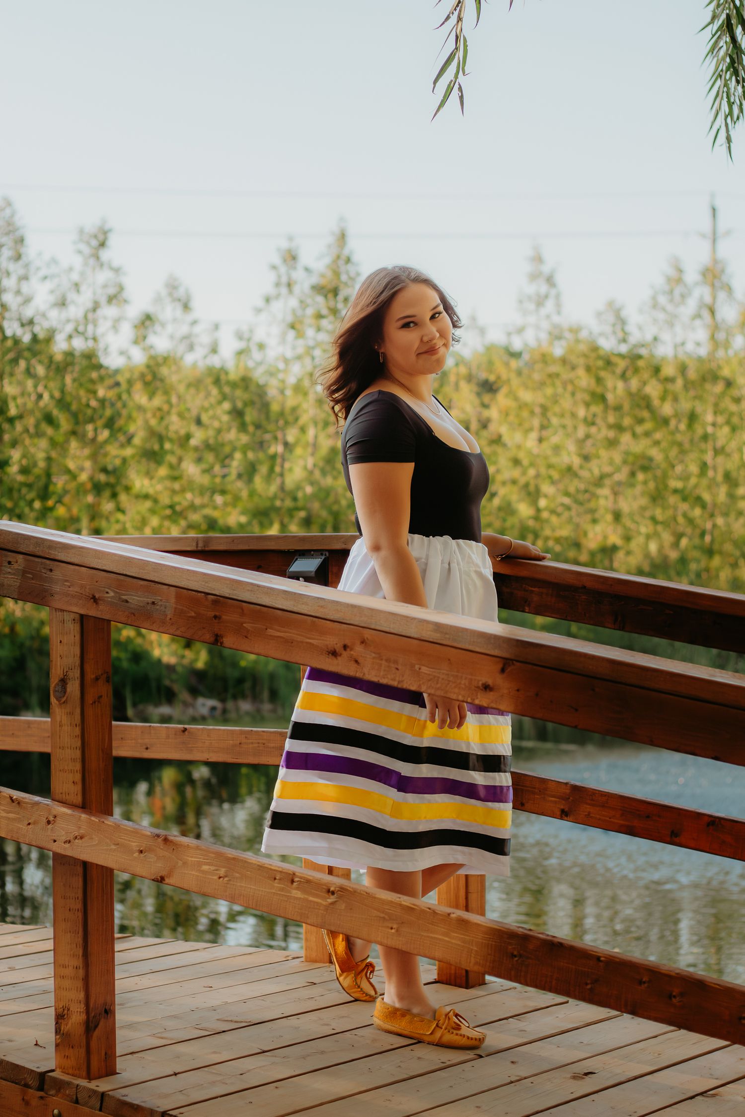 Graduate in a striped dress sits on wooden dock railing during golden hour.