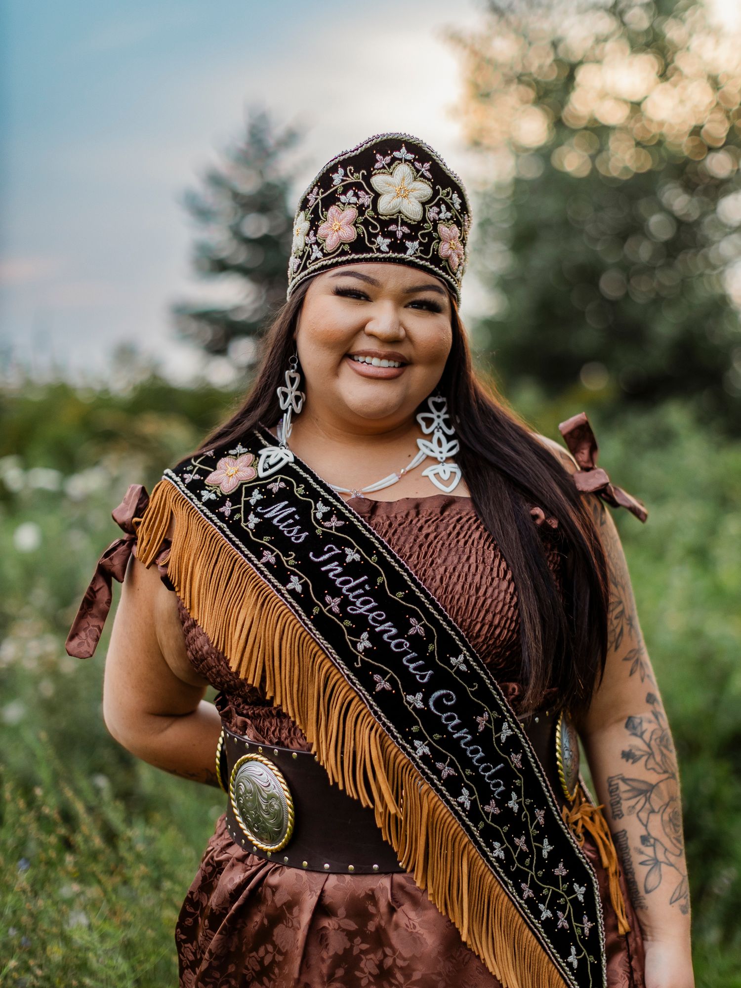 Miss Indigenous Canada titleholder smiling in traditional regalia, crown, fringe dress, and sash outdoors.