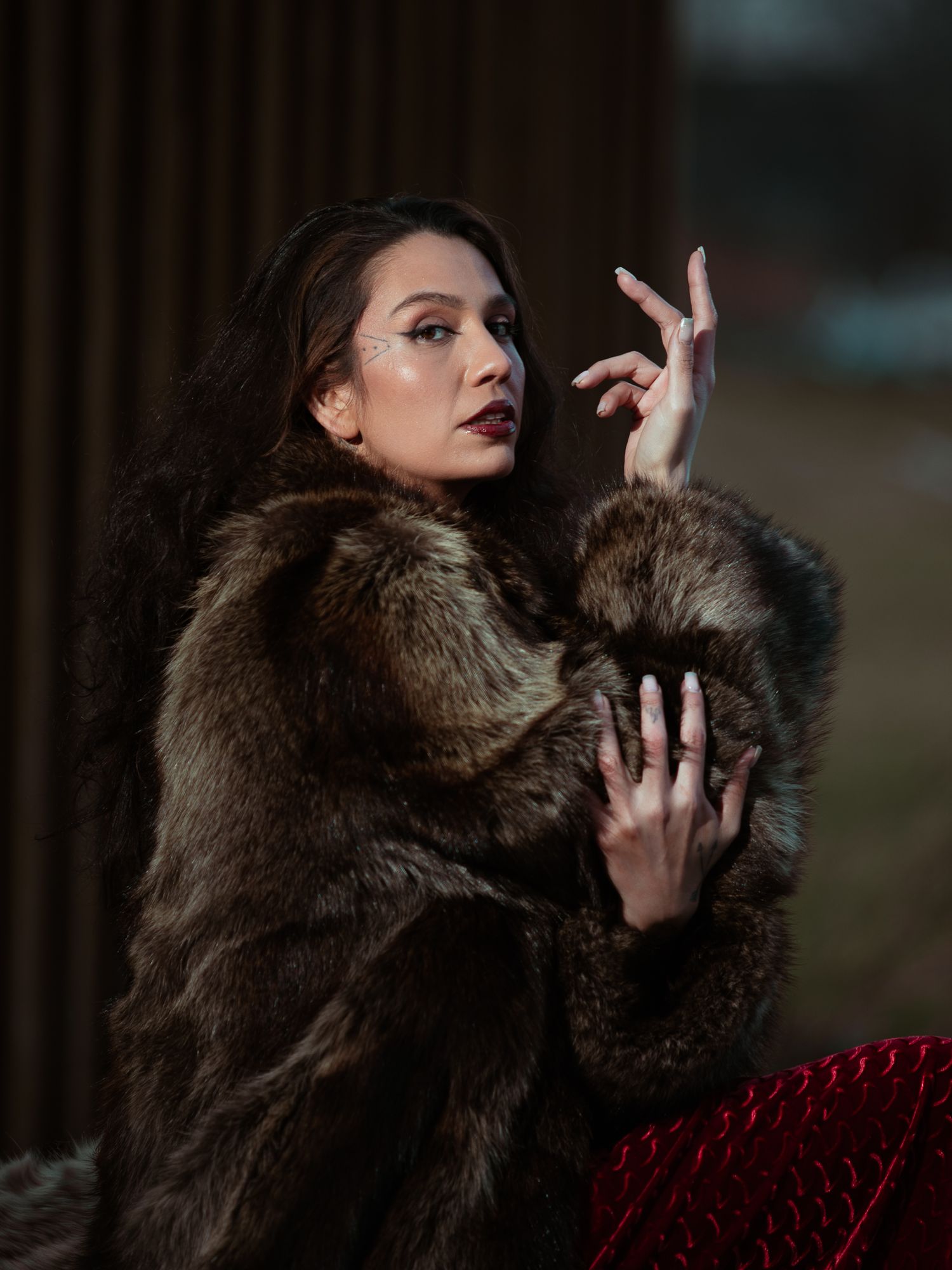 A woman in a luxurious brown fur coat poses elegantly with raised hands, wearing dramatic makeup and dark lipstick.