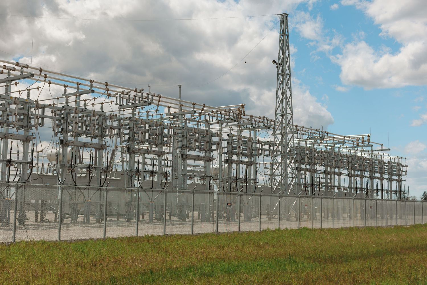 Large electrical substation with high-voltage transmission equipment surrounded by a security fence under cloudy skies.