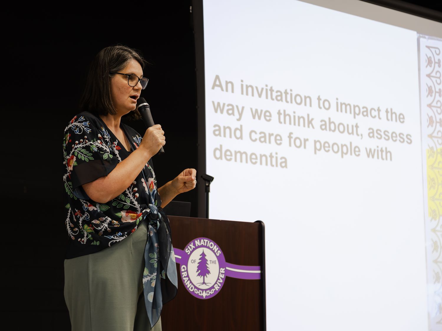 Woman speaks at dementia care conference podium beside slide about new approaches to assessing and caring for dementia.