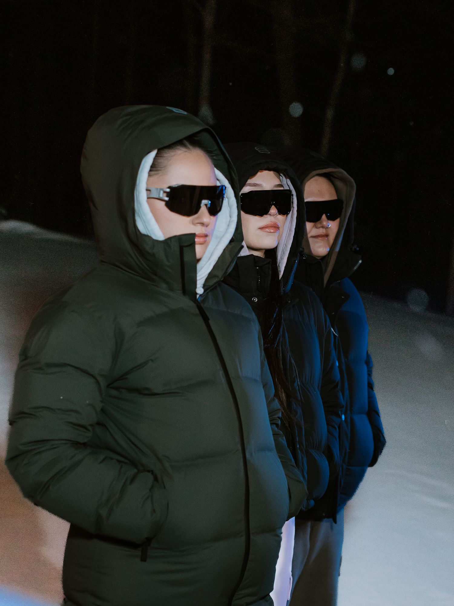 Three individuals in hooded puffer jackets and black sunglasses stand in a snowy winter landscape.