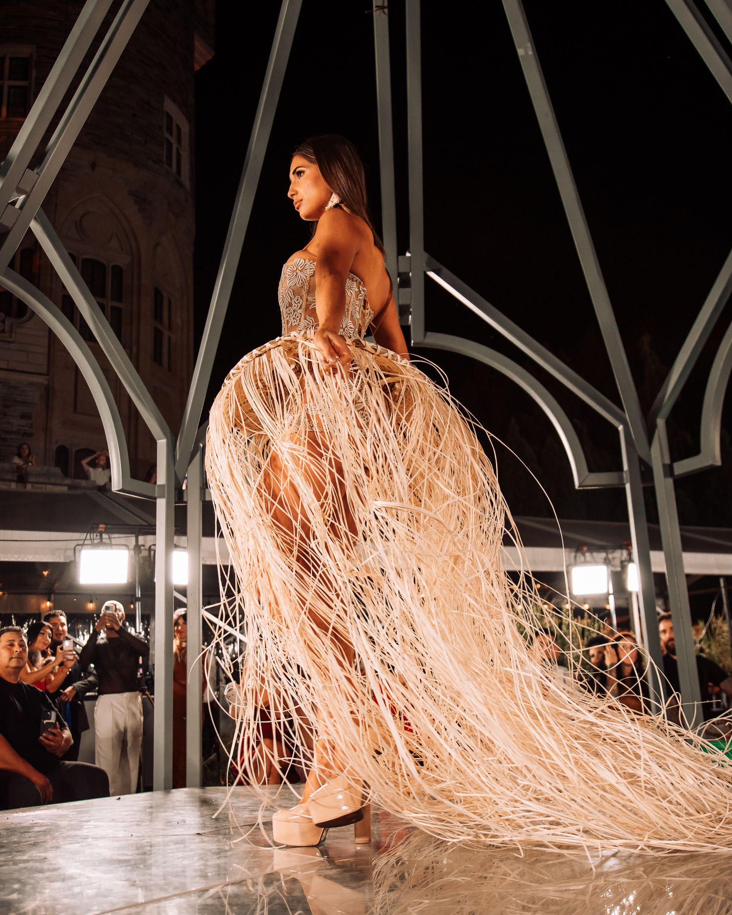 Model walks runway in ethereal beige fringe gown with dramatic flowing strands at night fashion show.