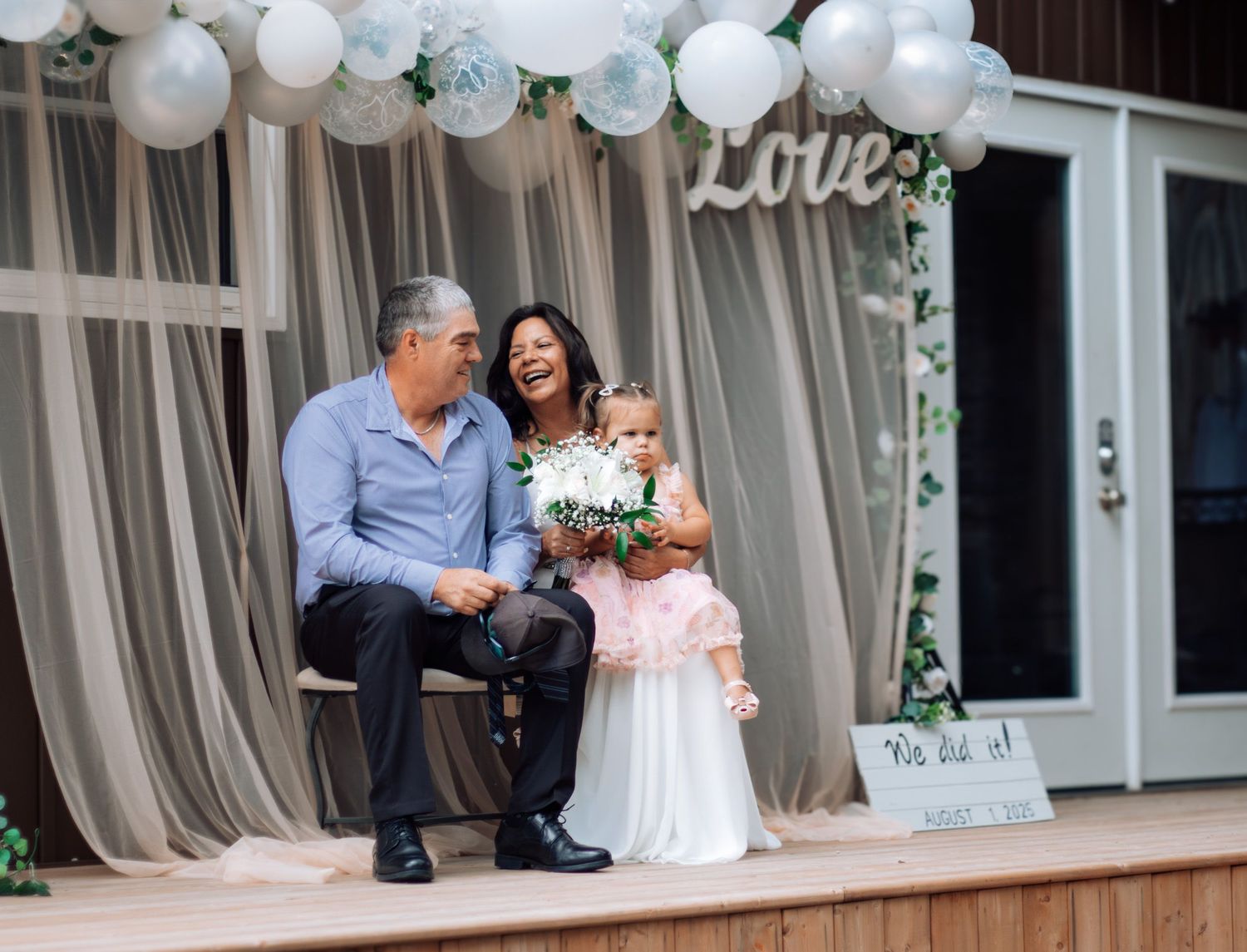 Bride and groom sit with young flower girl holding bouquet at decorated outdoor wedding reception.