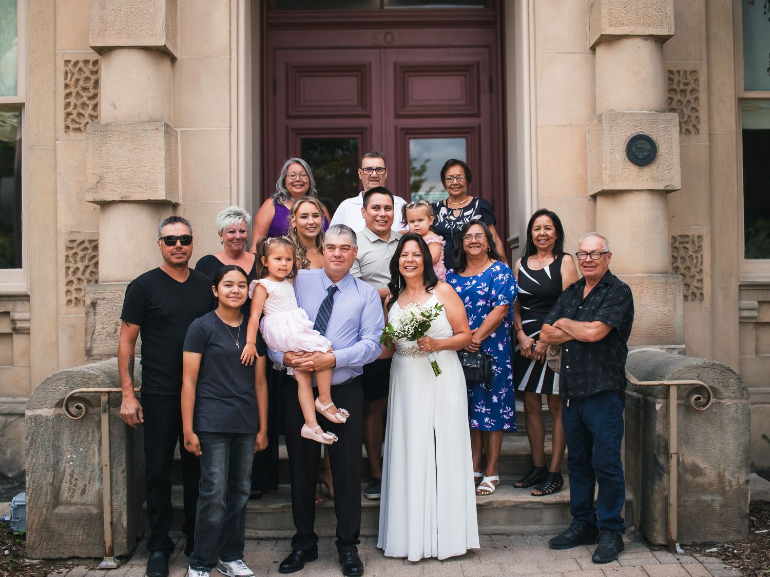 Wedding party group photo on stone building steps, bride in white gown holding bouquet with groom and family.