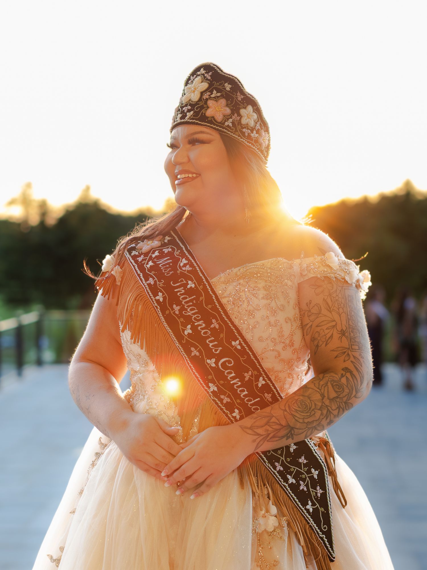 Woman in pageant crown and sash smiling outdoors at golden hour in elegant gown with tattoos.