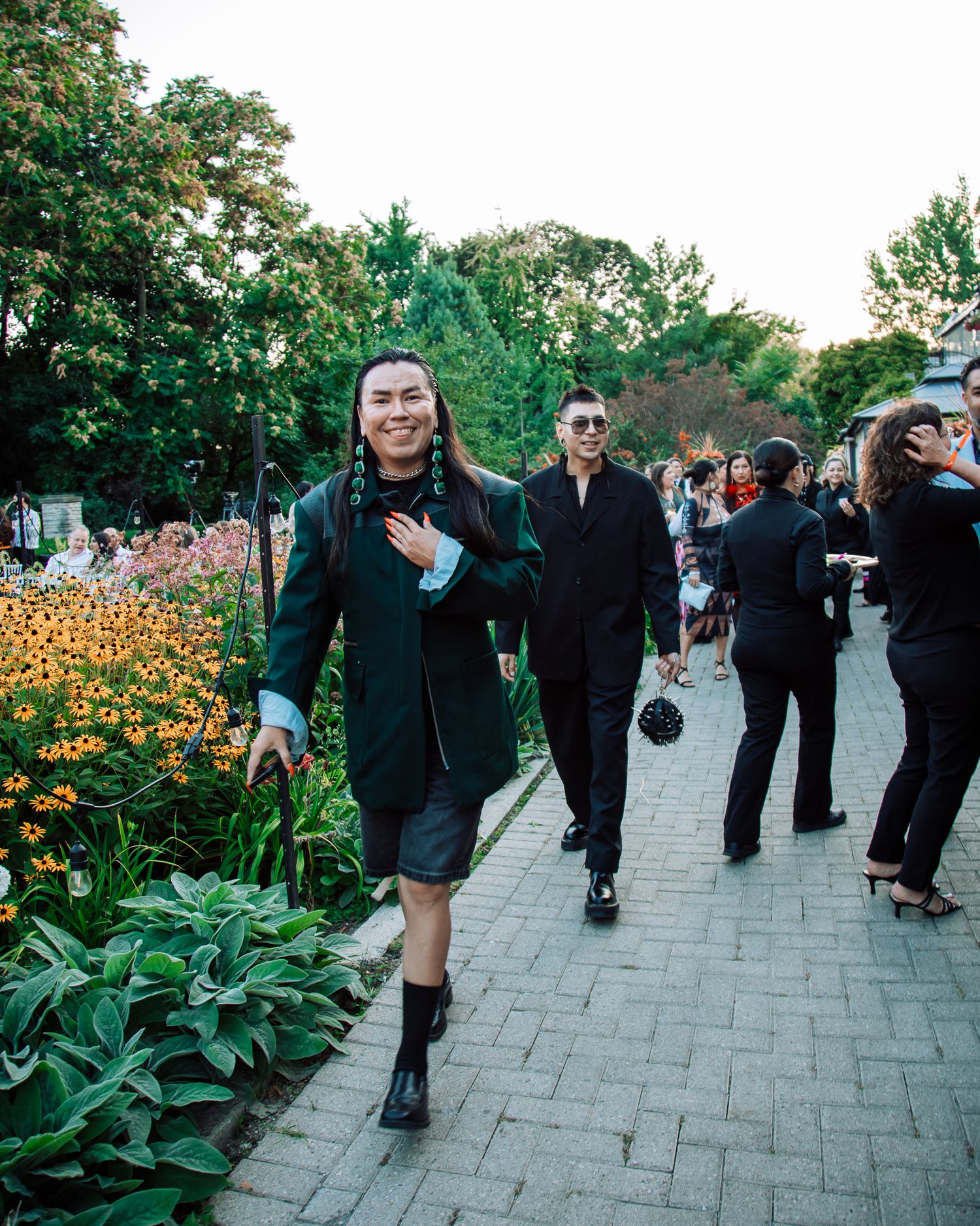 Smiling man with long hair walks garden path in black jacket at an outdoor fashion event.