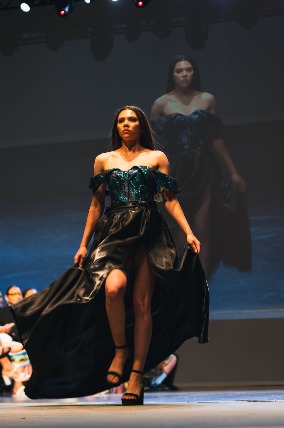 Model walks runway in black satin gown with emerald green sequin corset bodice at fashion show.