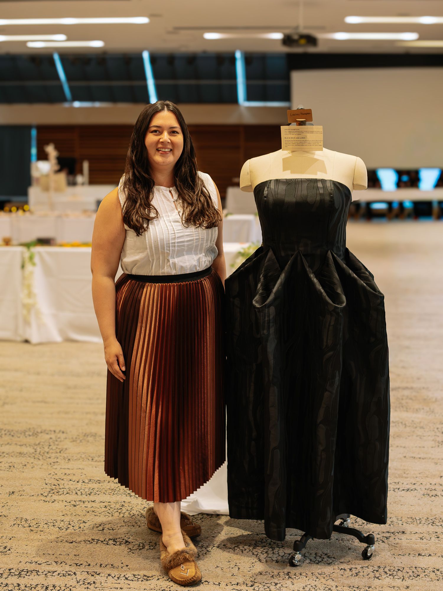 Woman in pleated skirt stands beside black strapless gown on mannequin at fashion showcase event.