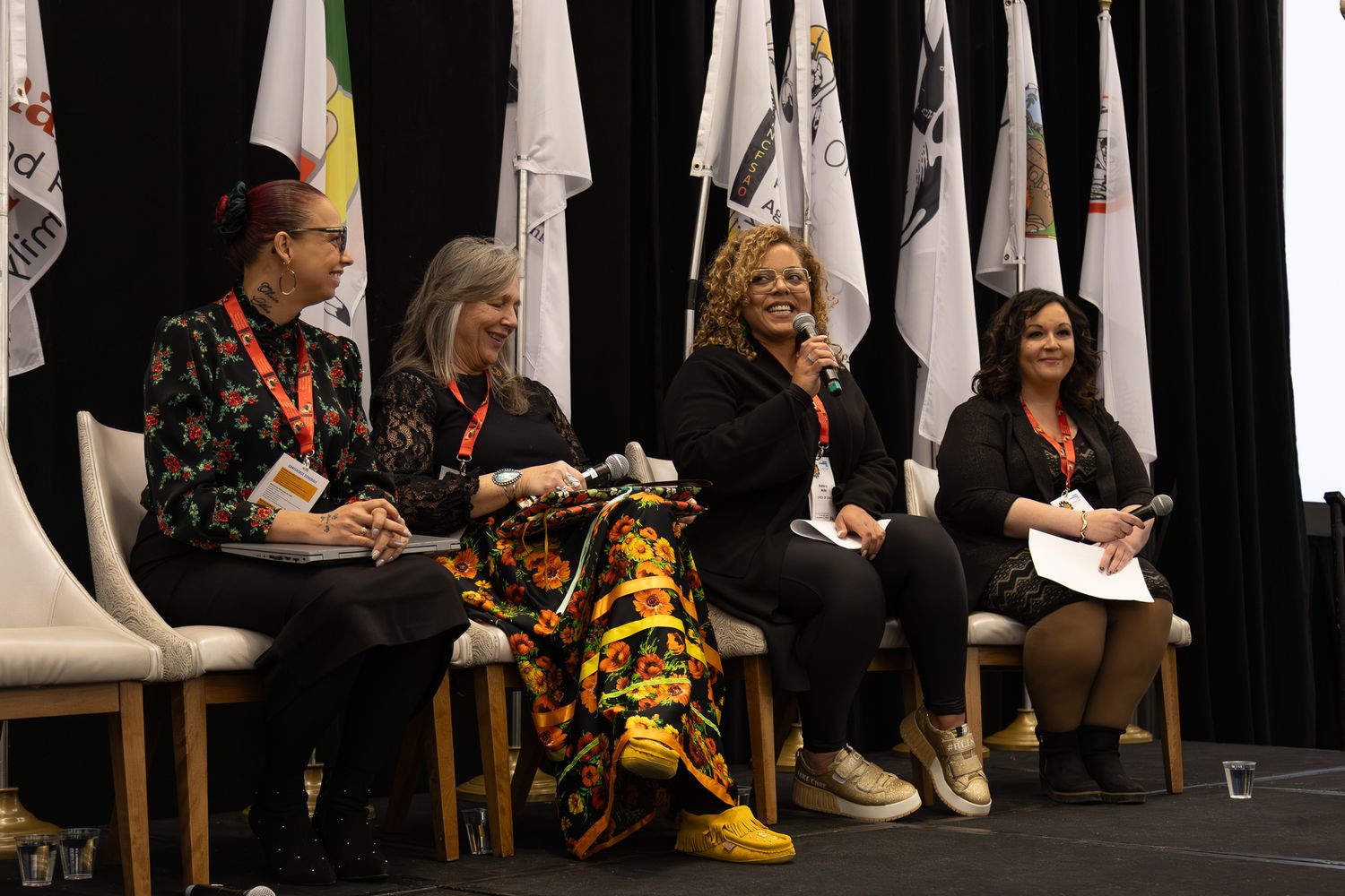 Four women seated on stage at a conference panel discussion with organizational flags displayed in the background.