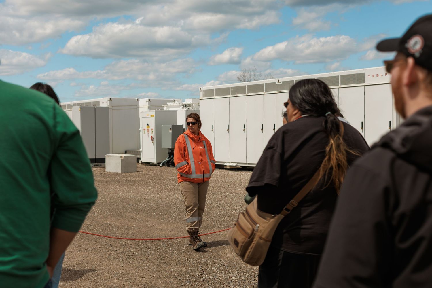 A worker in an orange vest presents to a group at an outdoor energy storage facility with large white battery units.
