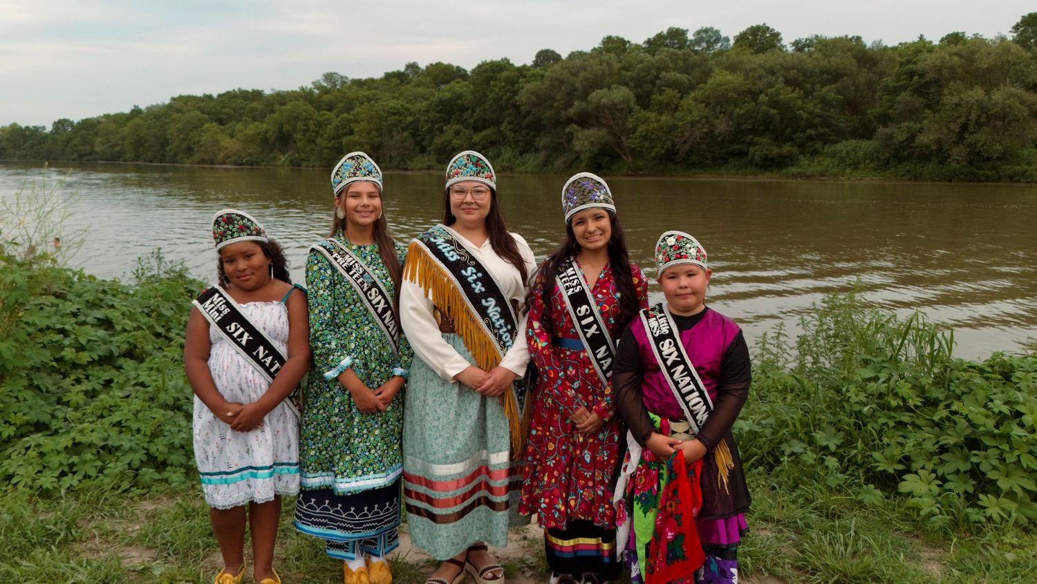 Five women in traditional Native American regalia and pageant sashes pose near a river with lush green trees in the background.